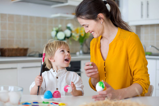 Beautiful blond child, toddler boy, painting easter eggs with mother at home, making easter wreath with rainbow colors - Valokuva, kuva