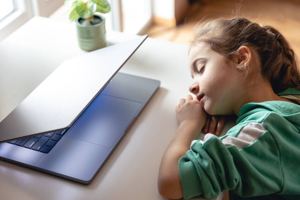 Pieni tyttö nukkuu kannettavan tietokoneen edessä pöydällä, väsynyt lapsi nukahti oppituntien aikana.. - Valokuva, kuva