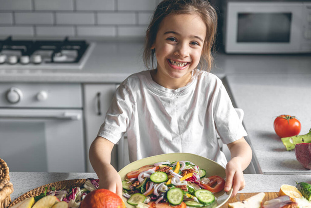 Little girl holding a plate of fresh vegetable salad while sitting in the kitchen, healthy eating concept. - Фото, зображення