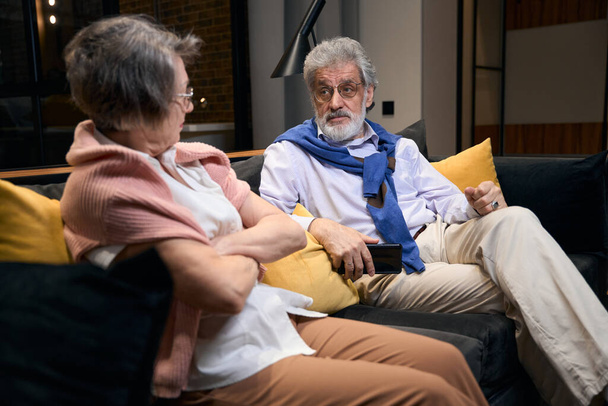 Bearded gray-haired man and elderly woman sort things out, the couple settled down on a sofa in a hotel room - Foto, Imagen