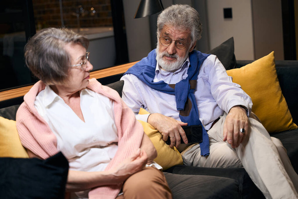 Bearded elderly man reproaches an upset woman, the spouses are located on a sofa in a recreation area - Photo, Image