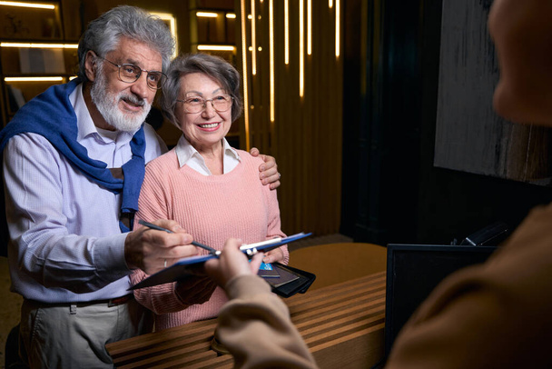 Traveling elderly couple checks into a hotel, the spouses register at the reception desk - Foto, Bild