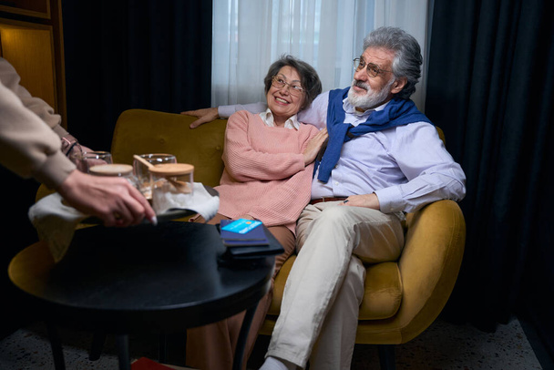 Charming grandparents relax in a hotel cafe, a man pays for drinks with a bank card - Foto, Imagen