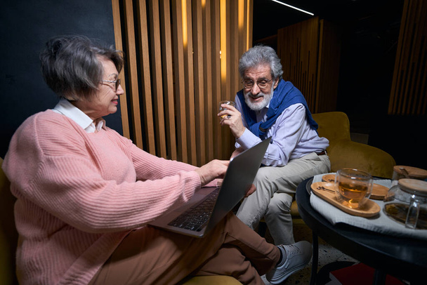 Pleasant mature couple chatting in hotel lounge area, woman has laptop on her lap - Fotó, kép