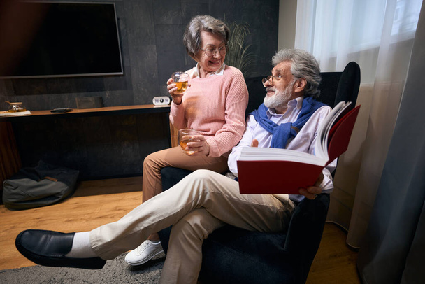 Old spouses are comfortably seated in an armchair with a book and drinks, people communicate pleasantly - Foto, afbeelding