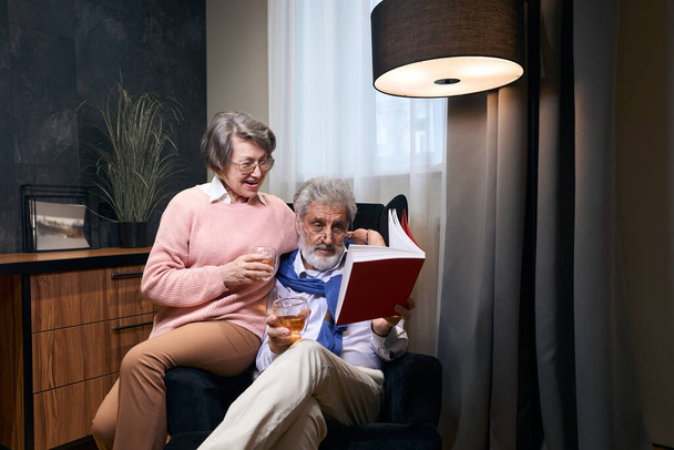 Cute grandparents settled down in an armchair with a book and drinks, people have a nice chat - Φωτογραφία, εικόνα