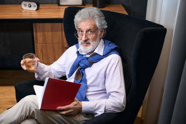Elderly man sits in an armchair by the window with a book and a drink, he has gray hair - Foto, Imagen