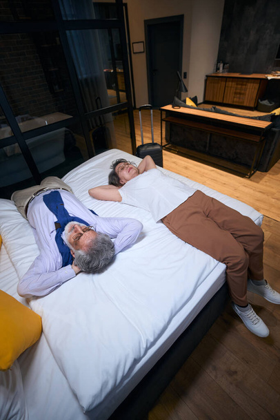 Moe van de weg, een oudere man en zijn metgezel rusten op een hotelbed, reiskoffer staat in de buurt - Foto, afbeelding
