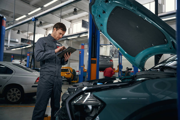 Mechanic in protective clothes standing in tire fitting near car with open hood and holding laptop - Foto, imagen