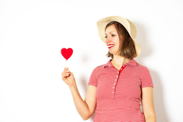 Retrato de la hermosa niña en camisa roja y sombrero de verano sobre un fondo blanco con una piruleta corazón rojo en la mano para el Día de San Valentín. Foto de alta calidad. - Foto, Imagen