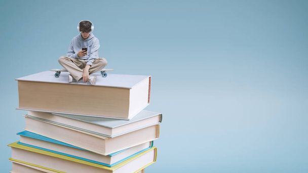 Young school student using a smartphone and sitting on a pile of books: online learning and education concept - Fotoğraf, Görsel