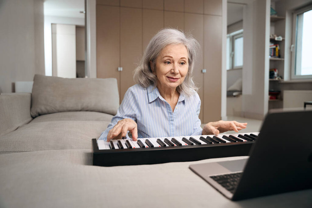 Pretty retired woman playing digital piano in living room, laptop standing in front of her - Fotó, kép