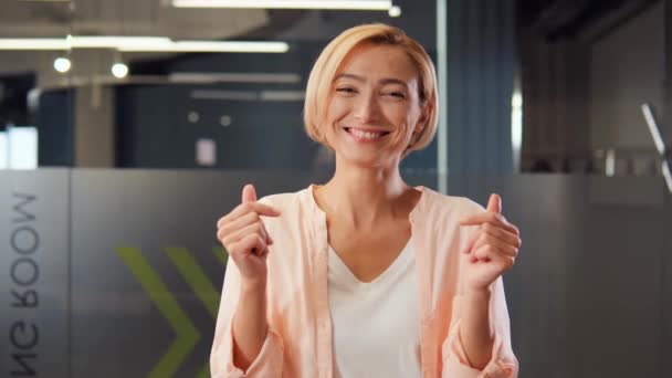 Het tonen van geld teken, slim en slim blond vrolijke vrouw met een sluwe glimlach in haar moderne kantoor geeft aan haar vermogen om geld te verdienen door het tonen van een cash gebaar en suggereren van grote winsten - Video
