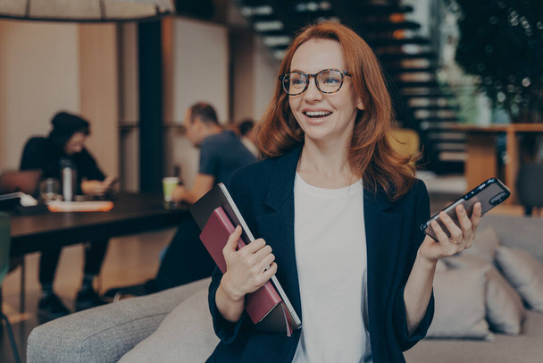 Smiling business woman with modern smartphone in hand, sitting on couch in lounge zone, holding laptop with notebook, smiling and looking around while waiting for meeting with client in office space - Photo, image