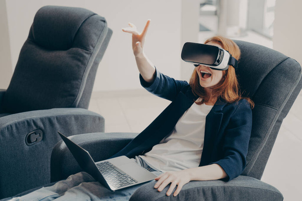 Sorpresa donna dai capelli rossi eccitato in VR occhiali auricolari e laptop, seduto in morbida poltrona grande in ufficio, cercando di toccare oggetti 3D realtà virtuale, puntando con indice in aria - Foto, immagini