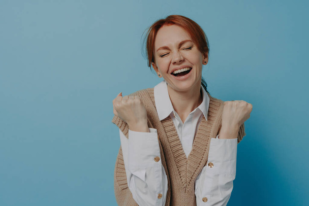 Young happy satisfied young red haired woman with closed eyes celebrating success, raising arms with clenched fists and screaming from happiness and excitement after achieving goal or job promotion - Photo, Image
