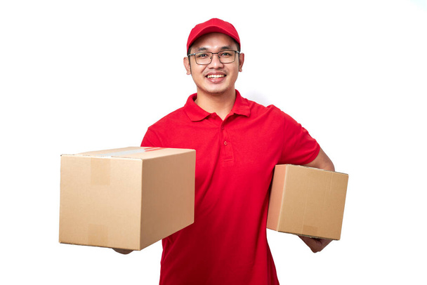 Smiling asian courier man wearing red uniform standing over white background holding two boxes with orders, packing shipping items for clients. - Fotó, kép