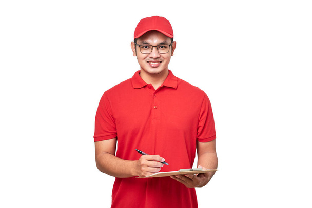 Νεαρός χαμογελαστός ασιάτης ντελιβεράς με κόκκινη στολή και καπάκι κρατώντας clip board σε απομονωμένο λευκό φόντο. - Φωτογραφία, εικόνα