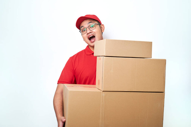 Sonriente feliz asiático repartidor hombre en rojo mensajero uniforme, mirando detrás de cajas con pedidos de clientes, llevar paquetes a casa del cliente aislado sobre fondo blanco - Foto, imagen