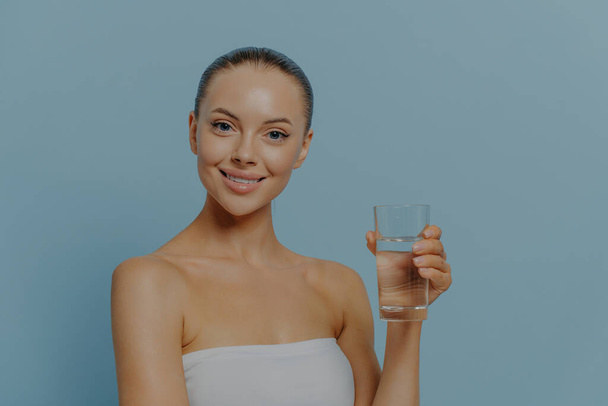 Schoonheid en gezonde levensstijl. Jonge vrolijke glimlachende goed uitziende vrouw die puur mineraalwater drinkt terwijl ze geïsoleerd boven een blauwe studio-achtergrond staat. Handhaving van vochtbalans en hydratatie - Foto, afbeelding