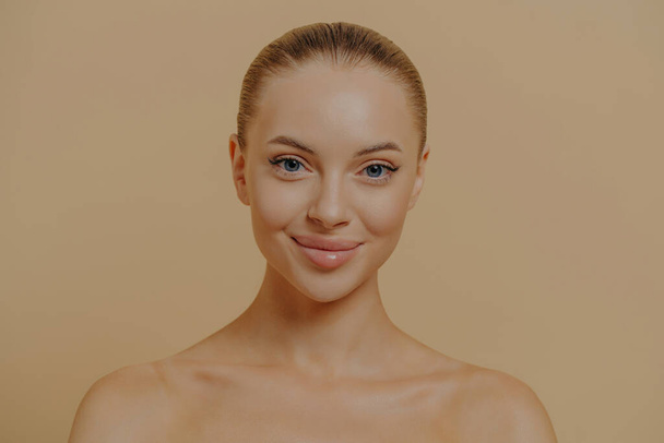 Beauty portrait of charming woman with healthy glowing skin posing seminude against beige wall, beautiful female face. Isolated shot of smiling female enjoying cosmetic skincare procedures - Fotografie, Obrázek