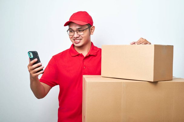 Asian courier man in red uniform smiling look at his smartphone while leaning oncardboard box parcel isolated over white background - Foto, afbeelding