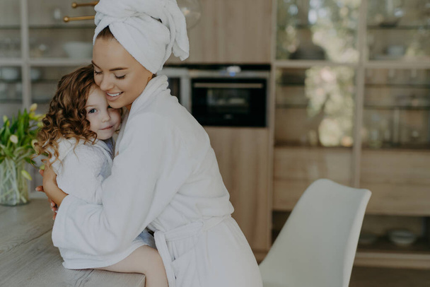 Happy affectionate young mother in bath towel and dressing gown embraces with love who sits at table feel refreshed after taking shower spend free time at home. Family beauty wellness concept - Photo, Image