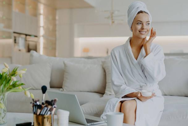 People beauty and skin care concept. Satisfied woman applies face cream enjoys facial treatments looks aside smiles gently uses cosmetic product poses at sofa in front of opened laptop computer - Zdjęcie, obraz