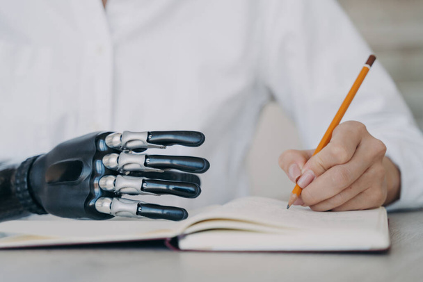 Close up of cyber arm and healthy limb on the notebook. Handicapped young woman with bionic prosthesis is studying remotely. Disabled girl is writing a book with pencil at the desk. Amputee lifestyle. - Фото, изображение