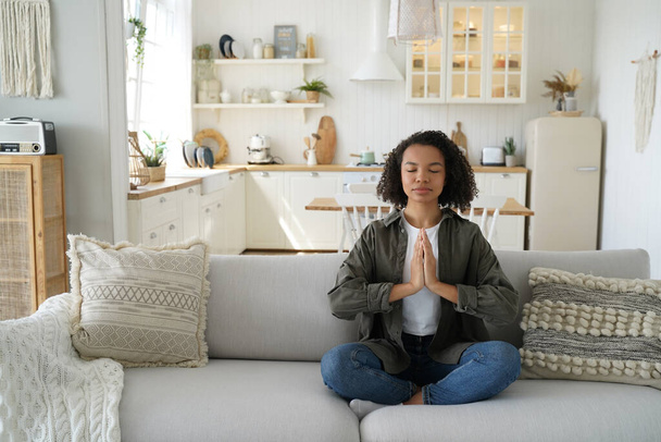 Curly jeune femme hispanique est assis sur le canapé dans la pose du lotus et la méditation. Adolescente afro fille pratique le yoga à la cuisine à la maison. Concentration et équilibre. Wellness et soulagement du stress. - Photo, image