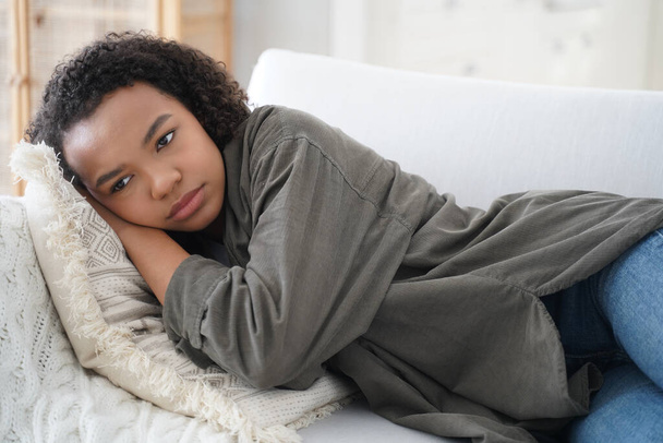 Απογοητευμένη μοναχική έφηβη είναι ξαπλωμένη στον καναπέ στο σπίτι. Η καταπιεσμένη Αφροαμερικανή έφηβη υποφέρει από εκφοβισμό ή βία. Ισπανίδα είναι απελπισμένη. Έννοια άγχους, άγχους και προβλημάτων υγείας. - Φωτογραφία, εικόνα