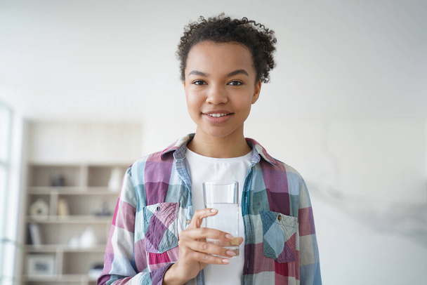 Glimlachend Afrikaans Amerikaans tienermeisje houdt glas vast met mineraalzuiver water dat thuis staat. Gelukkige jonge vrouw die naar de camera kijkt met vers zuiver aqua. Gezonde levensstijl, wellness concept. - Foto, afbeelding