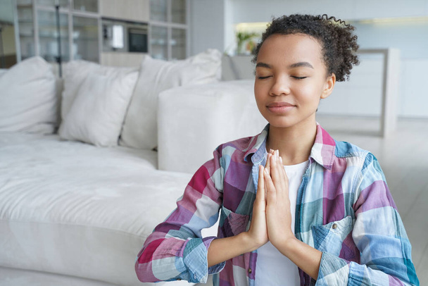 Spaanse jonge vrouw zit op de vloer in lotuspositie met haar ogen dicht. Tiener afro meisje is thuis yoga aan het oefenen. Rust en meditatie. Stressverlichting en concept van geestelijke gezondheid. - Foto, afbeelding