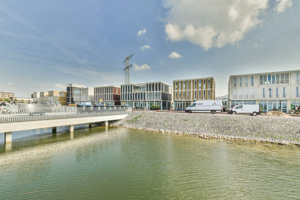 Amsterdão, Países Baixos - 10 de abril de 2021: uma ponte sobre a água com edifícios no fundo e carros estacionados do outro lado do rio abaixo - Foto, Imagem