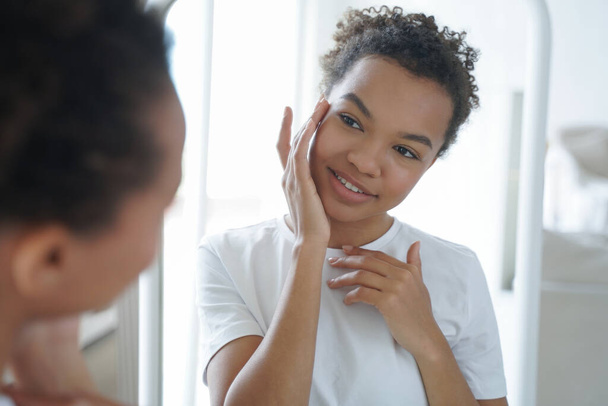 Mirror reflection of young attractive hispanic woman. Happy gorgeous african american girl applying cream to her face. Skin cleansing and treatment. Beauty routine and hygiene of tender teenage girl. - Foto, immagini