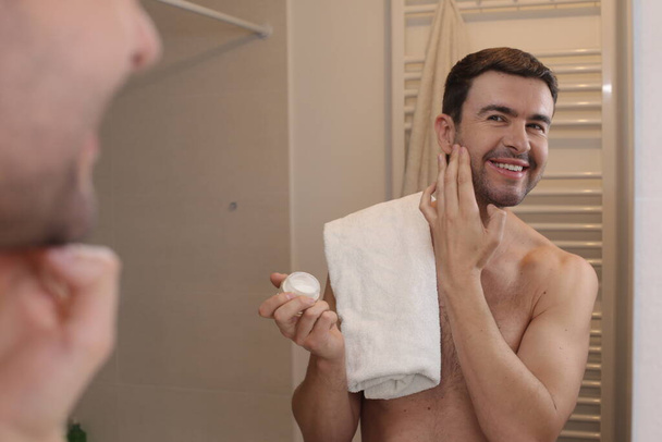 portrait of handsome young man applying beard balm in front of mirror in bathroom - Photo, Image