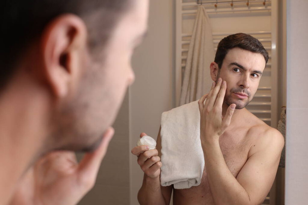 portret przystojnego młodego mężczyzny nakładającego balsam do brody przed lustrem w łazience - Zdjęcie, obraz