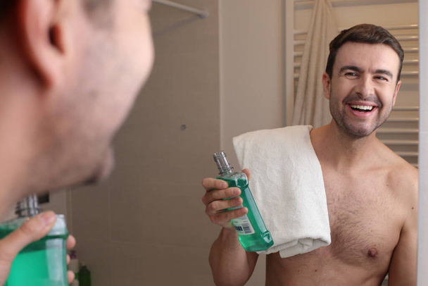 Porträt eines hübschen jungen Mannes mit Handtuch und Mundwasser vor dem Spiegel im Badezimmer - Foto, Bild