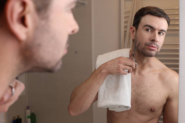 portrait of handsome young man cutting beard with scissors in front of mirror in bathroom - Photo, Image