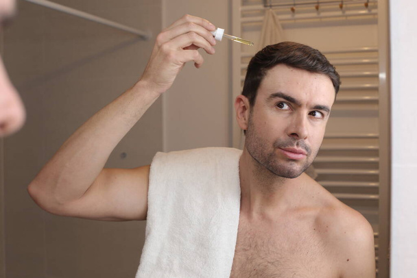 portrait of handsome young man applying hair oil in front of mirror in bathroom - Photo, Image