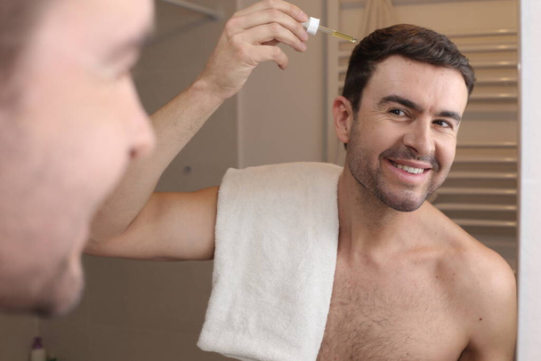 портрет красивого молодого человека, намазывающего волосы маслом перед зеркалом в ванной комнате - Фото, изображение