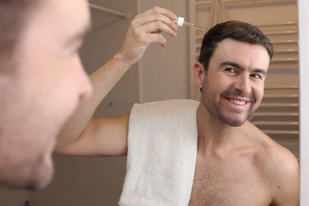 портрет красивого молодого чоловіка, який наносить масло для волосся перед дзеркалом у ванній
 - Фото, зображення