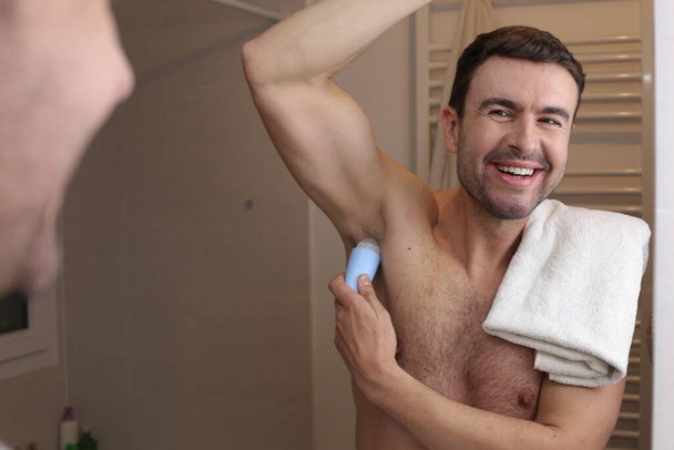 портрет красивого молодого человека, надевающего дезодорант на подмышки перед зеркалом в ванной комнате - Фото, изображение