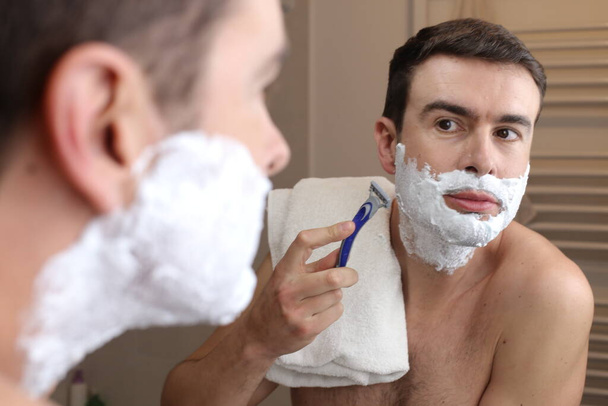 портрет красивого молодого человека, бреющегося перед зеркалом в ванной комнате - Фото, изображение