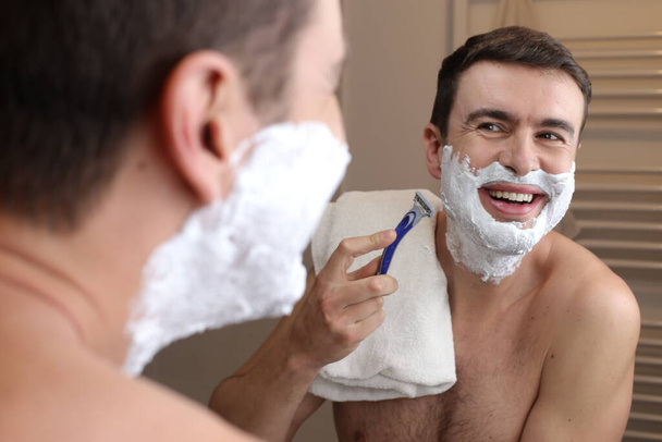 portrait of handsome young man shaving in front of mirror in bathroom - Zdjęcie, obraz