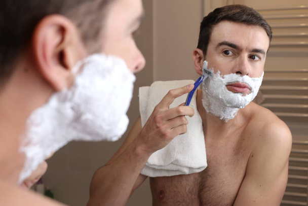 portrét pohledného mladého muže, jak se holí před zrcadlem v koupelně - Fotografie, Obrázek