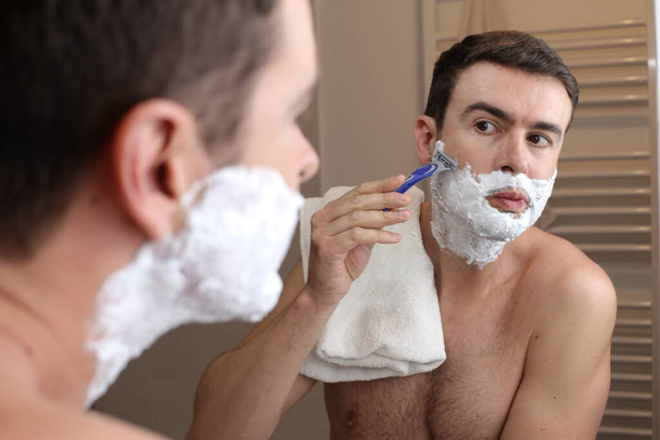 портрет красивого молодого человека, бреющегося перед зеркалом в ванной комнате - Фото, изображение