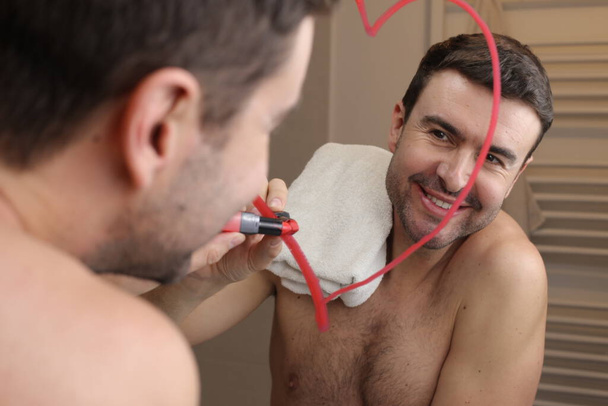 portret van knappe jongeman in de voorkant van spiegel in de badkamer, tekenen hart met lippenstift - Foto, afbeelding