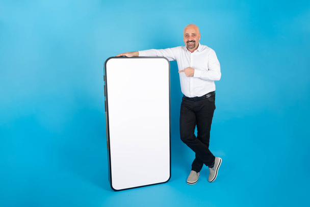 Big huge smartphone, full body length of happy man leaning big huge smartphone. Caucasian man pointing empty white blank screen for mock up. Mobile app advertisement concept idea. Blue background. - Foto, immagini