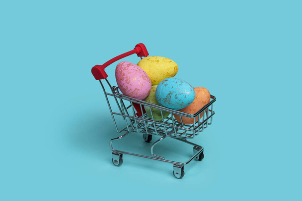 Un carrello pieno di uova di Pasqua colorate decorate con motivi divertenti e vivaci su uno sfondo blu pastello. Perfetto per promozioni pasquali, biglietti di auguri e post sui social media. - Foto, immagini